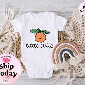 May include: White baby onesie with a cute orange illustration and the text "little cutie".