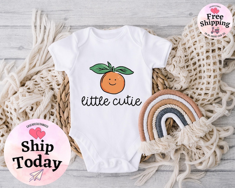 May include: White baby onesie with a cute orange illustration and the text "little cutie".