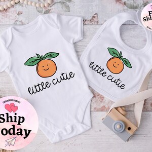 May include: White baby bodysuit and bib with a smiling orange graphic and the text "little cutie".