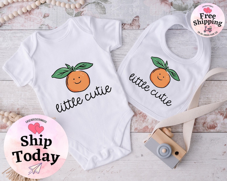 May include: White baby bodysuit and bib with a smiling orange graphic and the text "little cutie".