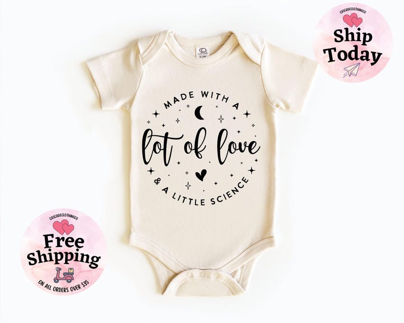 May include: Cream-colored baby onesie with short sleeves. The front features a circular design with the text "MADE WITH A lot of love & A LITTLE SCIENCE" in black script. The design includes stars, a crescent moon, and a heart.