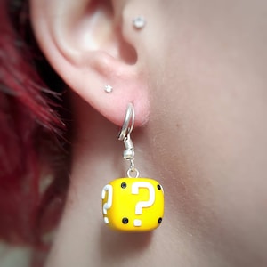 以下が含まれることがあります： 黒いドットが付いた黄色のクエスチョンマークブロックのイヤリング。イヤリングは銀色のフックにぶら下がっています。