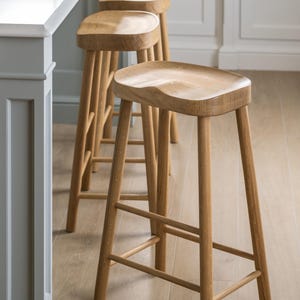 May include: Wooden bar stools with a light brown finish. The stools have a curved seat and four legs with a footrest. The stools are lined up in front of a white countertop.
