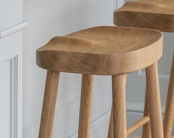 May include: Two bar stools crafted from light-toned oak wood, featuring four tapered legs, horizontal support rungs, and a contoured, saddle-shaped seat with a visible wood grain pattern, positioned in a pair for elevated seating.