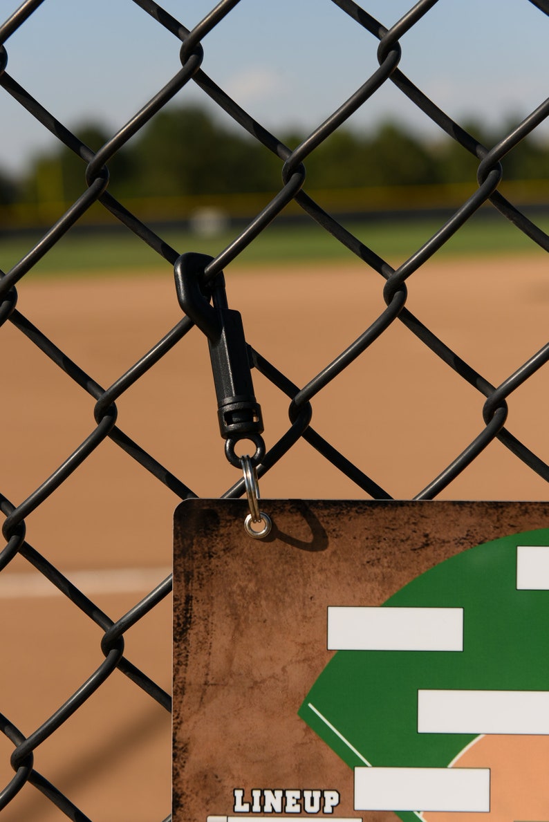 May include: A baseball lineup card hanging on a chain-link fence. The card has a brown background with a green baseball field graphic and white boxes for player names. A black carabiner clip attaches the card to the fence.