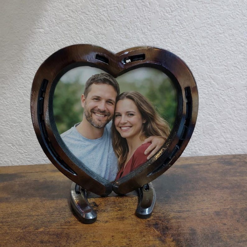 May include: A heart-shaped photo frame made from horseshoes. The frame holds a photo of a smiling couple. The frame is brown with a glossy finish and sits on two horseshoes. The background is a light-colored wall and a wooden surface.