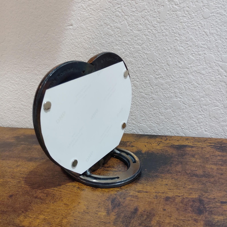 May include: A heart-shaped photo frame with a horseshoe base. The frame is black with a white insert held in place by four small magnets. The base is a silver horseshoe. The frame is displayed on a wooden surface.