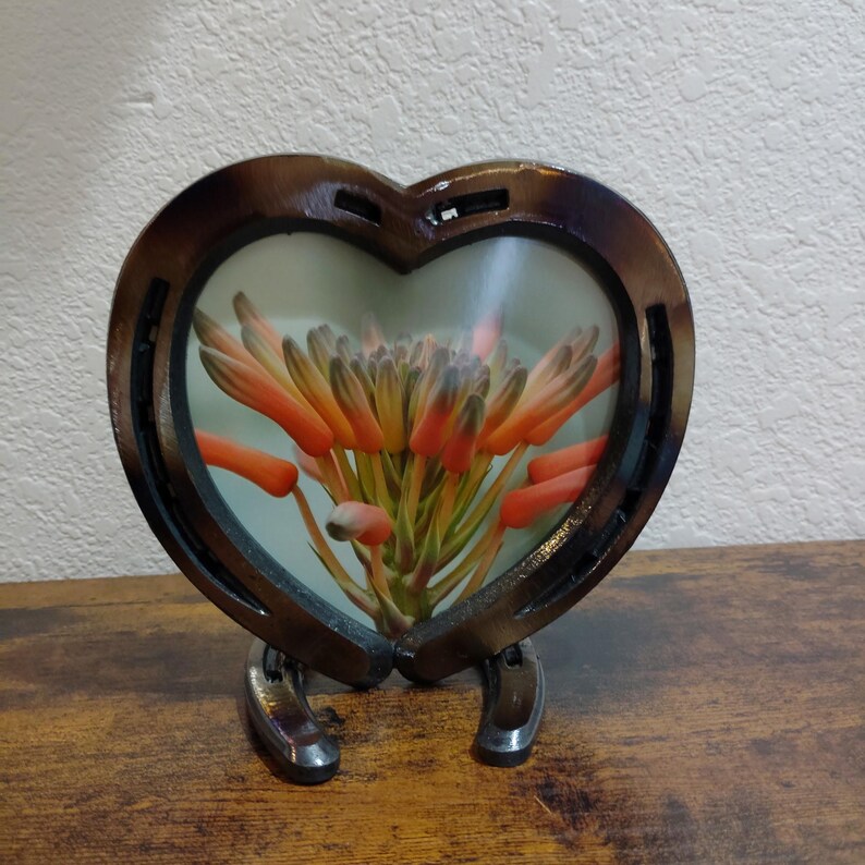 May include: A heart-shaped frame made from a horseshoe, holding a floral arrangement. The frame is brown with a glossy finish, and the flowers are orange and green. The frame is supported by two horseshoes.