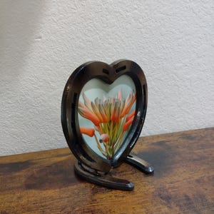 May include: A heart-shaped picture frame made from horseshoes. The frame holds a floral arrangement with orange and pink blooms. The frame is supported by two horseshoes, creating a unique decorative piece.