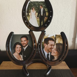 Könnte beinhalten: Ein rustikaler Metall-Hufeisen-Fotorahmen mit drei Hochzeitsfotos. Der Rahmen besteht aus dunklem Metall und hat ein einzigartiges Hufeisen-Design. Jedes Foto zeigt ein Paar in Hochzeitskleidung.