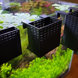 Op de afbeelding: Drie zwarte plastic plantenmanden met een rasterpatroon zijn bevestigd aan de zijkant van een glazen aquarium. De manden zijn leeg en klaar om gevuld te worden met planten.
