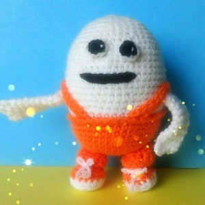 Puede incluir: Un personaje blanco de crochet con forma de huevo que lleva un mono naranja y zapatos blancos. El personaje tiene ojos negros y una boca negra.