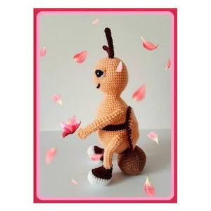 May include: A crocheted plush toy in the shape of a brown and orange insect with a round body and long legs. The toy is sitting on a white surface with pink flower petals scattered around it.
