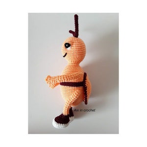 May include: A crocheted orange and brown bee toy with a black eye. The bee is wearing brown shoes and has a brown stripe around its middle. The text "Aix in crochet!" is visible in the image.
