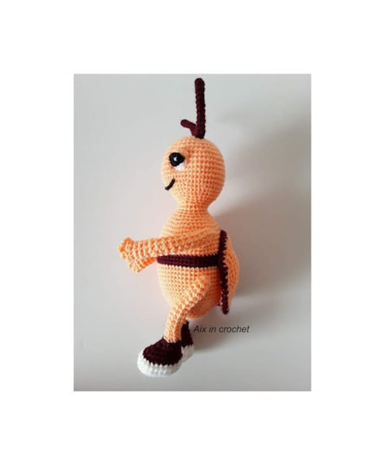 May include: A crocheted orange and brown bee toy with a black eye. The bee is wearing brown shoes and has a brown stripe around its middle. The text "Aix in crochet!" is visible in the image.