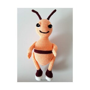 May include: A crocheted bee plush toy with orange and brown yarn. The bee has a smiling face with black eyes and a black mouth. The bee has white feet.