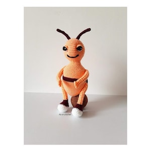 May include: A crocheted bee toy with orange and brown stripes, sitting on a brown ball. The bee has white eyes and a small smile.