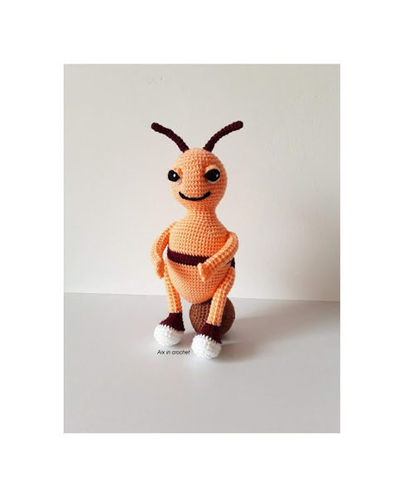 May include: A crocheted bee toy with orange and brown stripes, sitting on a brown ball. The bee has white eyes and a small smile.
