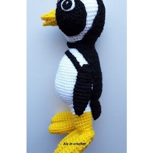 Op de afbeelding: Een gehaakt pingu&iuml;n speelgoed met zwart-witte strepen, een gele snavel en gele voeten. De pingu&iuml;n staat op een wit oppervlak. De tekst "Aix in crochet" is zichtbaar onderaan de afbeelding.