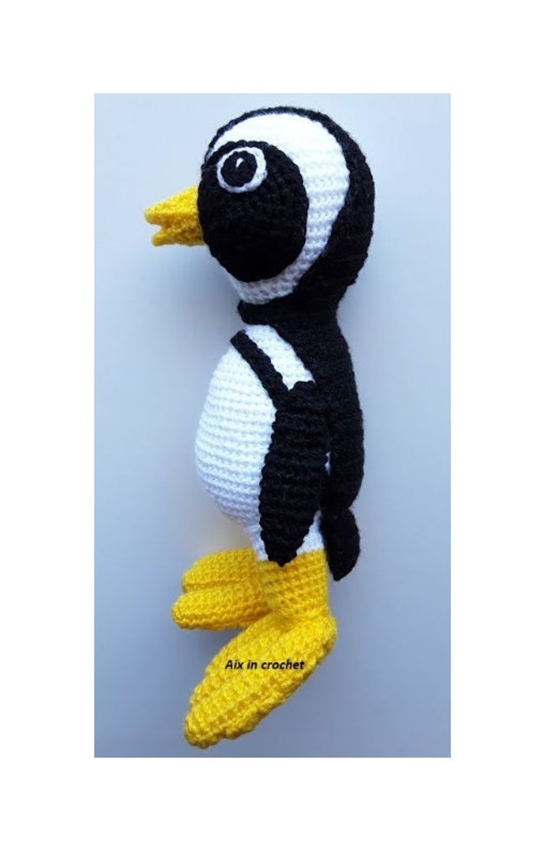 Op de afbeelding: Een gehaakt pingu&iuml;n speelgoed met zwart-witte strepen, een gele snavel en gele voeten. De pingu&iuml;n staat op een wit oppervlak. De tekst "Aix in crochet" is zichtbaar onderaan de afbeelding.