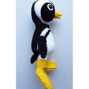 Op de afbeelding: Een gehaakt pingu&iuml;n speelgoed met zwart-witte strepen, een gele snavel en gele voeten. Het speelgoed staat op een wit oppervlak. De tekst "Aix in crochet" is zichtbaar op de voet van het speelgoed.
