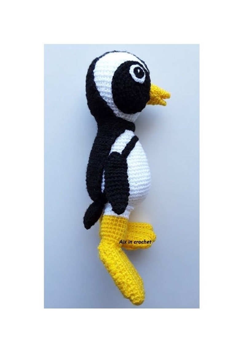 Op de afbeelding: Een gehaakt pingu&iuml;n speelgoed met zwart-witte strepen, een gele snavel en gele voeten. Het speelgoed staat op een wit oppervlak. De tekst "Aix in crochet" is zichtbaar op de voet van het speelgoed.