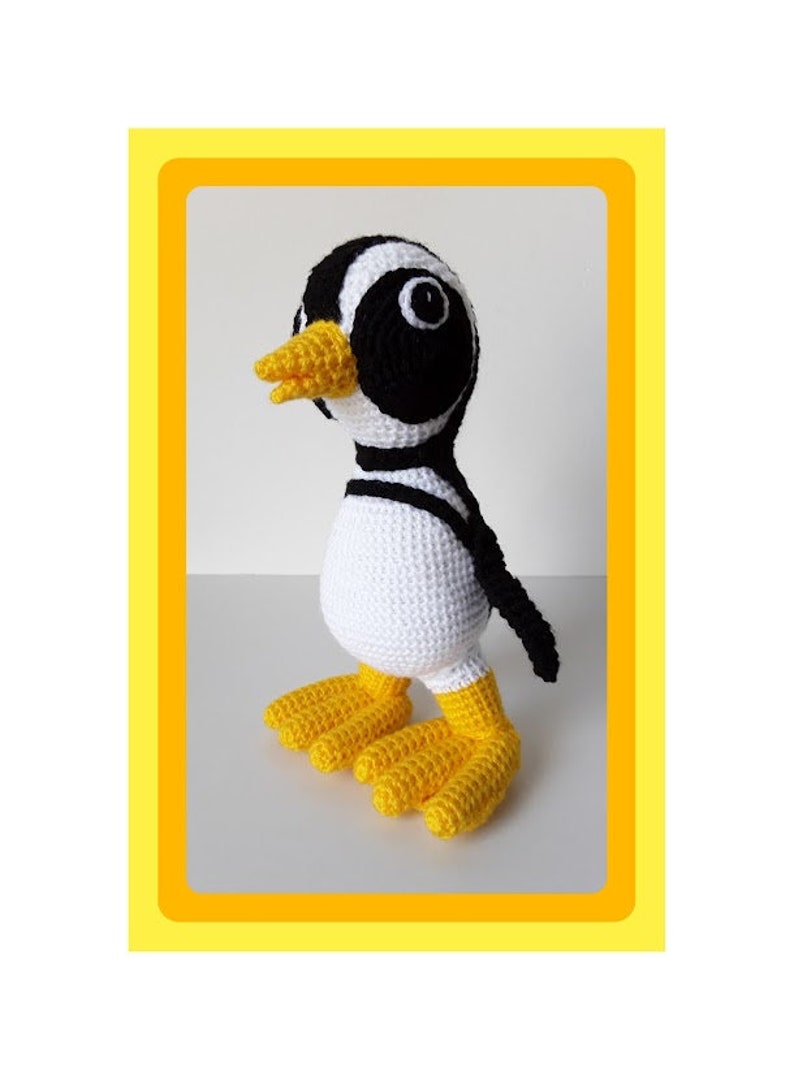 Op de afbeelding: Een gehaakt pingu&iuml;n speelgoed met zwart-witte strepen, een gele snavel en gele voeten. De pingu&iuml;n staat op een wit oppervlak.