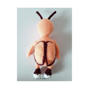 May include: A crocheted stuffed toy shaped like a bee with a peach-colored body, brown antennae, and white shoes. The bee is standing on a white surface.