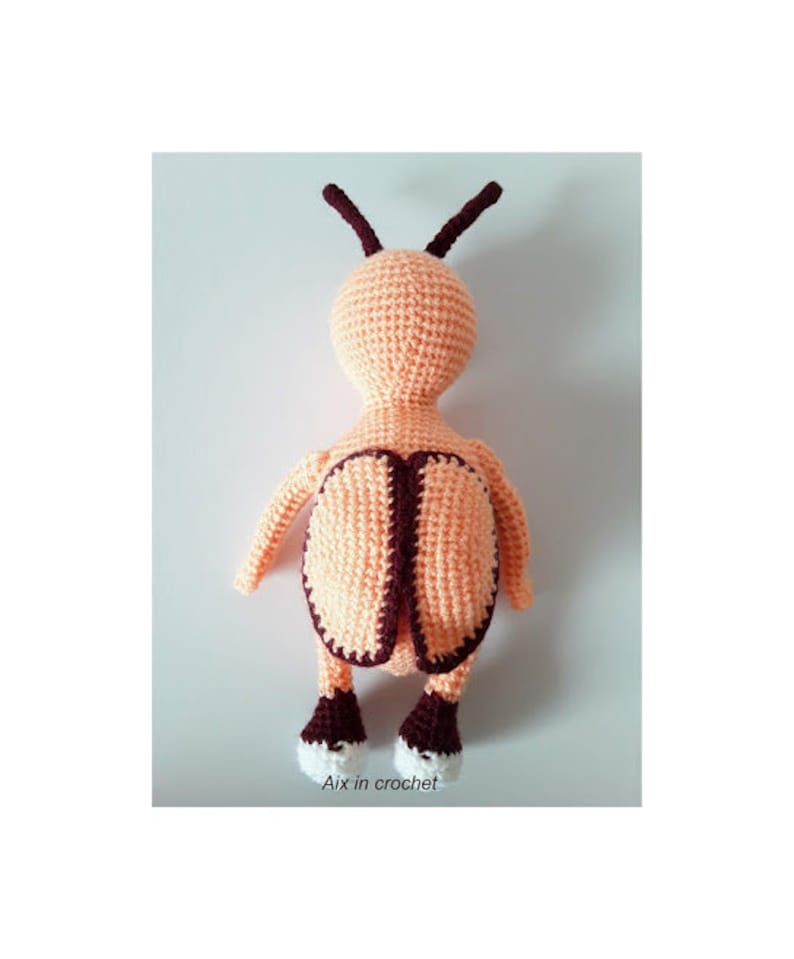 May include: A crocheted stuffed toy shaped like a bee with a peach-colored body, brown antennae, and white shoes. The bee is standing on a white surface.