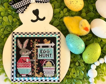 May include: White wooden bunny cutout with a black and white checkered bow. The bunny is holding a cross-stitch piece with a bunny in a red sweater and the words "Egg Hunt". The bunny is surrounded by green foliage and white beads.