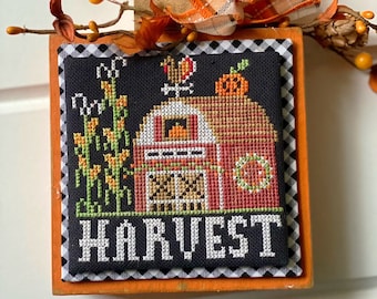 May include: A framed cross-stitch design with a black and white checked border. The design features a red barn with a pumpkin, a rooster, and corn stalks. The text "HARVEST" is written below the barn.