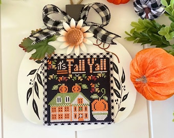 May include: A white pumpkin-shaped wooden sign with a stitched design that reads "It's Fall Y'all." The sign is decorated with a black and white checkered bow, a metal sunflower, and small orange and white pumpkins. Green foliage surrounds the sign.