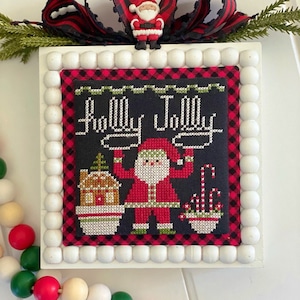 Pode incluir: Um bordado de ponto cruz enquadrado em branco com fundo preto e borda de xadrez vermelho e preto. O bordado representa o Papai Noel segurando um bastão de caramelo, uma casa de gengibre e uma tigela de bastões de caramelo. O texto "Holly Jolly" está bordado em branco sobre o Papai Noel.