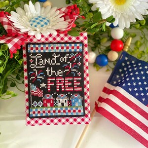 May include: A red, white, and blue patriotic cross-stitch with the text "Land of the Free" surrounded by a red and white checkered fabric. A small American flag is on a wooden stick to the right of the cross-stitch.