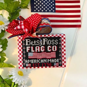 May include: A red, white, and blue gingham fabric square with a black background featuring the text "Betsy Ross Flag Co. American Made Est. 1776". The square is decorated with a red, white, and blue bow.