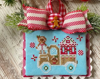 May include: A cross-stitched Christmas ornament featuring a light blue background with a gingerbread truck design. The ornament is framed with a red and white scalloped edge and topped with a red and white striped bow. A red and white bead garland is in the background.