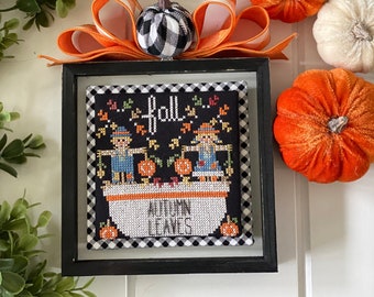 May include: A black framed cross-stitch with a black and white checked border. The cross-stitch depicts two scarecrows in a bathtub with pumpkins and the text "...fall..." and "Autumn Leaves".
