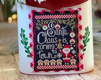 May include: A white metal mailbox with a red trim and a red and black buffalo plaid fabric with a white trim. The mailbox has a cross-stitch design on the front that says "So Santa Claus is coming to town".