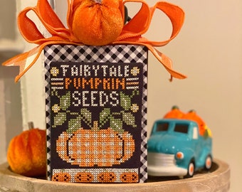 May include: A tiered wooden tray featuring a black and white checked box with the text "Fairytale Pumpkin Seeds" and a cross-stitch image of a pumpkin. The box is adorned with a large orange velvet pumpkin and a white and orange bow. A blue toy truck with a bed full of orange pumpkins sits on the top tier of the tray. The bottom tier displays a white velvet pumpkin, an orange velvet pumpkin, and a green plant.