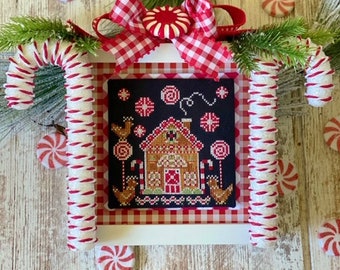 May include: A cross-stitch pattern featuring a gingerbread house with candy canes and a red and white gingham border. The pattern is framed with white and red candy cane trim.