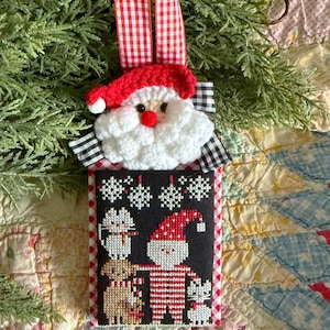 Può includere: Un ornamento natalizio fatto a mano con una testa di Babbo Natale all'uncinetto e un nastro a scacchi rosso e bianco. L'ornamento rettangolare mostra un disegno a punto croce di Babbo Natale, un cane e due gatti, con fiocchi di neve su sfondo nero.