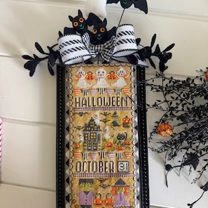May include: A rectangular framed cross-stitch sampler featuring Halloween motifs, including a haunted house, ghosts, a black cat, and the text "HALLOWEEN OCTOBER 31". The frame is topped with a black and white striped bow, felt bats, and a ghost.