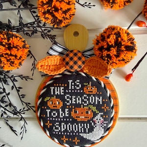 May include: A round wooden plaque featuring a black cross-stitch fabric insert with the text 'Tis the season to be spooky' surrounded by orange pumpkins and a ghost. It is decorated with a black and white striped bow, orange pom-poms, and leaves.