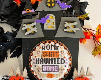 May include: A black and gray wooden Halloween house with a white cross-stitched sign that reads "Home Sweet Haunted". The house has a purple witch's hat, a purple bat, and a white picket fence. There are two orange pumpkins with black faces at the bottom of the house.