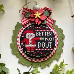 May include: A red and white checkered fabric ornament with a green felt leaf border. The ornament features a cross-stitch design of Santa Claus with the text "Better Not Pout".