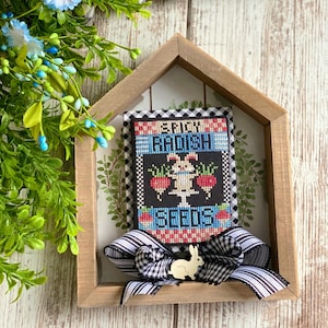 May include: A framed cross-stitch design featuring a bunny holding a basket of red radishes with the text "Spring Radish Seeds". The frame is made of wood and has a black and white checkered ribbon tied around it.