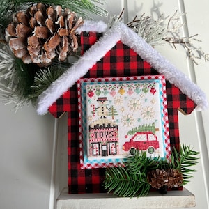 Puede incluir: Un adorno en forma de casa de cuadros rojos y negros con ribete de piel sintética blanca. La casa presenta una pequeña escena de punto de cruz de un coche rojo con un árbol de Navidad en la parte superior, pasando por una tienda con un muñeco de nieve en frente.
