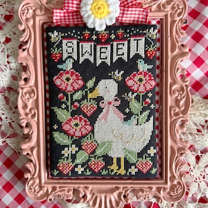 May include: A framed cross-stitch featuring a white duck with a pink bow, surrounded by red flowers and strawberries. The word "SWEET" is stitched across the top. The frame is pink, with a daisy and red and white gingham ribbon.