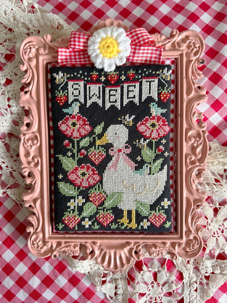 May include: A framed cross-stitch featuring a white duck with a pink bow, surrounded by red flowers and strawberries. The word "SWEET" is stitched across the top. The frame is pink, with a daisy and red and white gingham ribbon.