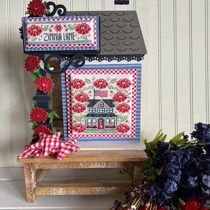 May include: A decorative wooden house-shaped sign with a cross-stitch design. The sign features the text "Zinnia Lane" and a cross-stitched house with red flowers and an American flag. The sign is decorated with red paper flowers.